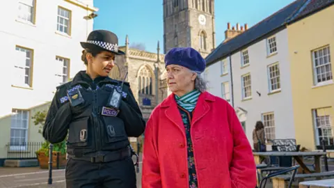 Two women look at each other in a street: A police officer in uniform and an older woman in a red coat and beret.