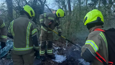 Fire crews in protective equipment look at the embers of a fire in woodland.
