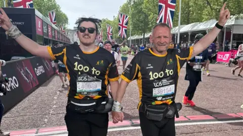 Beau and Gary McKee hold hands as they cross the finish line at the London Marathon. They are both wearing black and yellow tops that read: "100 to LDN". Their free hands are raised in the air. There are union jack flags erected down the Mall behind them.