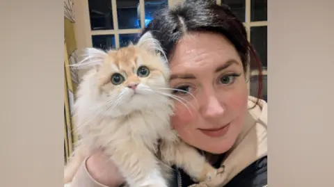 The Cat Care Co Sheree George has dark hair in a pony tail and is wearing a beige hooded top and black jacket and is holding a white fluffy cat and smiling at the camera