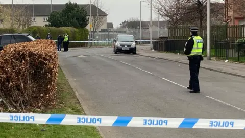 Police officers on a road, a section of which has been cordoned off with blue and white police tape at either end