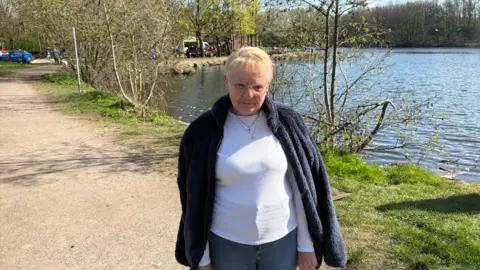 LDRS A woman with short blonde hair and wearing glasses, a white long-sleeved top and a blue fleece, is standing on a footpath by the side of the lake.