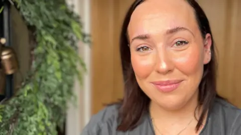 Laura Foster/BBC Carys Davies with long dark hair, wearing a grey T-shirt. She is in a house with a wooden door behind her and a holly wreath to her right.