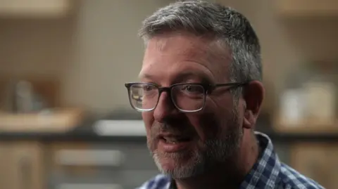 Frank Tipping wears glasses and a blue and white checked shirt, pictured from the shoulders up. He has gray hair and a thick beard. The background of the image is blurred. 