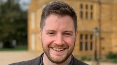 Richard Clinton, facing the camera, smiling. He is wearing a blue shirt, with a brown jacket and is standing in front of a building, which is blurred. 