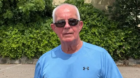 A mostly bald man with a bit of grey hair wearing sunglasses and a blue t-shirt standing in front of a hedge in a car park.