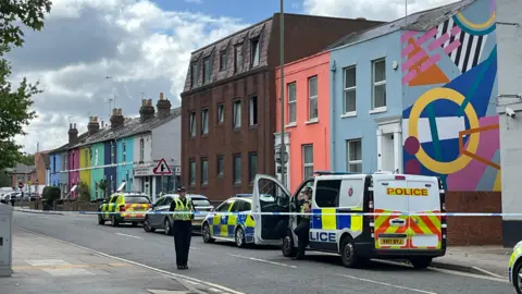 Police tape cordons off a road in Gloucester, which has colourfully painted houses dotted along it. There are a number of police vehicles - including a van - parked along it. A police officer is stood behind the cordon, surveying the surroundings, while another climbs out of the passenger door of the van.