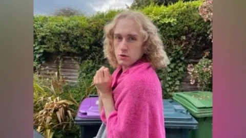 A person with shoulder-length curly hair is pictured in a garden wearing a pink top.