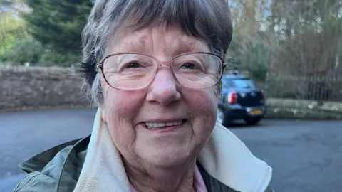 An older woman with short greying hair and glasses smiles while wearing a green and cream coat standing in a car park with a black car behind her.