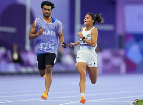 Getty images Simran and Abhay are running together. He is on the right side of the picture and he is on the left. Their bodies are reflecting each other so that both his right leg and his left leg is thrown out. They are both catching their tithters. He is wearing a white sleeveless top and shorts; He is wearing a blue sleeveless top with the word "Guide" Written and black shorts on it. Both have orange running shoes. 