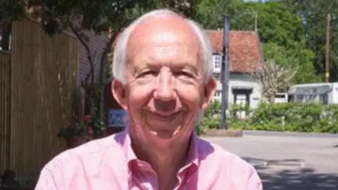 Neil Watts smiles at the camera while standing outside. He wears a pink shirt and has grey hair.