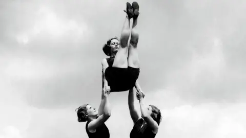 A black and white image of two women holding a third woman up in the air while an acrobatic exercise. They are wearing black leotards. The woman at the top of the image has her legs up in the air. 