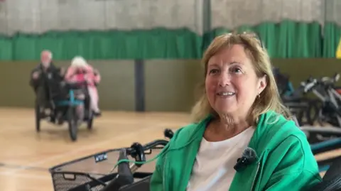 Caroline sits to the right of the image on one of the tandem bikes. She has shoulder length hair wearing a green zip-up hoodie and a white t-shirt. She's looking off to the right of the camera.
In the background there is a the selection of bikes. To the right there is a couple who are on a tandem side by side bike blurred out.