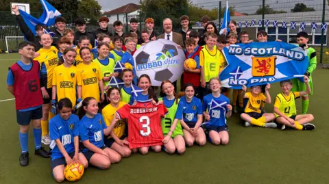 A group of schoolchildren in football strips  on a pitch. They are holding a Scotland top with Andy Robertson's name and a football with East Robbo-shire written on it.