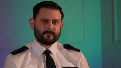 PC Jack Taylor, in police uniform, is sitting in a room lit with blue and green lights