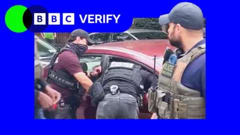 BBC Um policial se inclina pela janela esmagada de um carro vermelho para prender um homem enquanto outros dois assistem.