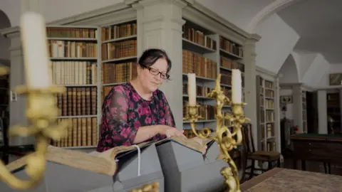 Longleat Emma Challinor in the library of Longleat looking over a large, old volume. She wears a floral top and glasses.