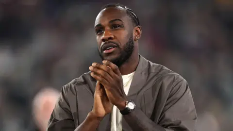 A man standing on a pitch with a blurred purple light in the background. He has black hair in cornrows and a closely trimmed moustache and beard. He is smiling and looking into the distance. He is wearing a brown bomber jacket, cream t shirt and numerous gold and silver necklaces with charms.