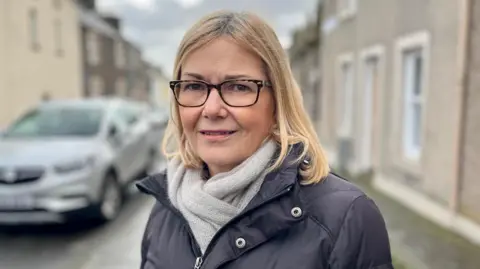 Wendy Copeland has shoulder length blonde hair, glasses, a grey scarf and a dark jacket. She is standing in a street.