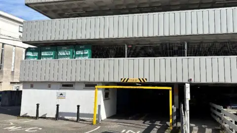 The ground level entrance to the car park has a yellow height restriction barrier and road markings saying In and M C, standing for motor cycles. Two more storeys can be seen above withy grey walls.