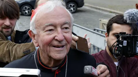 Cardinal Vincent Nichols has white hair and is wearing a red skull cap, black soutane with red trimmings, waistband and buttons as he is pursued by journalists on his way to a meeting with fellow cardinals in the Vatican