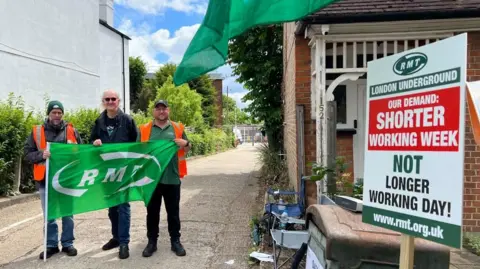 Three people stand on a residential street holding a green RMT union flag, with a large sign reading “London Underground – Our demand: shorter working week, not longer working day".