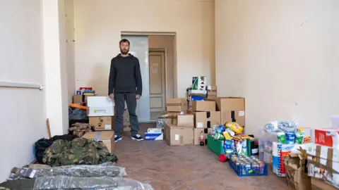 ADAM READ Chris stands in a room filled with humanitarian supplies