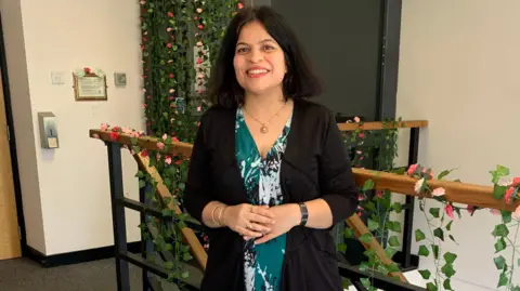 An Indian woman stands at the top of a staircase which is decorated with trailing green leaves and pink flowers. She is wearing a dark green dress with a white pattern and a black cardigan. she has dark hair in a long bob and is smiling into the camera.