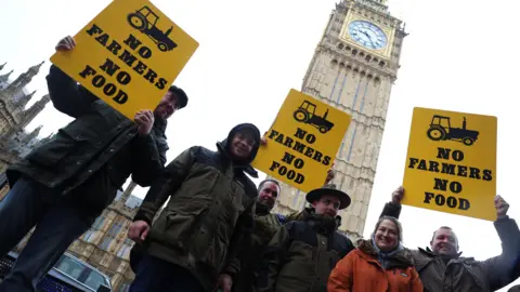 Getty Images A group of farmers holding yellow signs that say 'no farmers no food' in black stand outside Big Ben. 