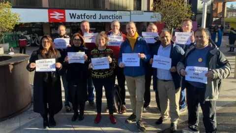 A group of 11 men and women outside Watford Junction railway station, holding small A4 paper signs which say "FAIR FARES FOR WATFORD", "REZONE WATFORD STATIONS" and "SIGN THE PETITION NOW". Most are wearing raincoats. The Watford Junction station sign is visible in the background.