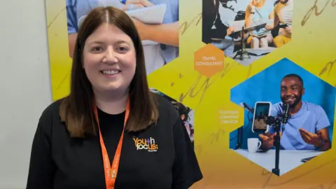 George Walker/BBC A young woman wearing a black t-shirt and bright orange lanyard. She is standing against a wall with different pictures of career options on it with a yellow background. She is smiling.