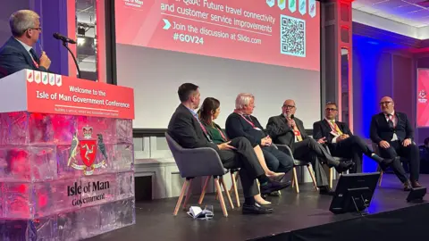 Five men and a women sit in a semi circle on chairs on a stage. One is speaking into a microphone. There are large red signs in the background with the Government Conference branding and another man is standing by a lectern holding a pen to his mouth.