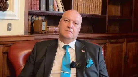 BBC A bald man sitting in a dark red chair in front of dark wood panelling. He is wearing a suit with a white shirt and blue time. He has a union jack pinned to his lapel.