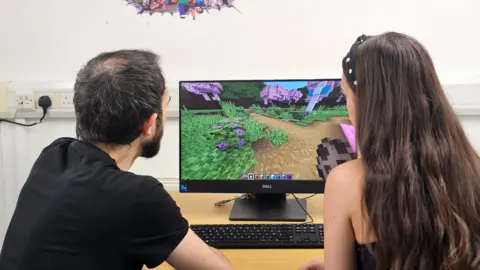 A man and a woman sitting at a desk with a desktop on it. They are playing Minecraft.