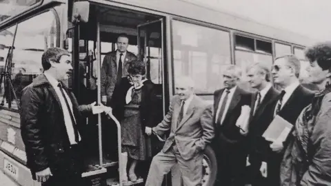 Kenny Beckers/Wales News Service A black and white photo of a bus, with Kenny Beckers in a white shirt and black tie chatting to six men and a woman