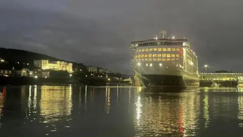 The Manxman - a large white-and-black ship - photographed in the middle of Douglas Harbour at night. Bright lights are reflected in the dark and calm harbour waters.