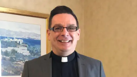 Justin Kennedy A man wearing rectangle glasses smiles at the camera, the picture is cropped to just his head and shoulders but you can see he is wearing a clerical collar - better known as a dog collar - and a grey blazer. The background shows yellow wallpaper and a painting of a Greek town.