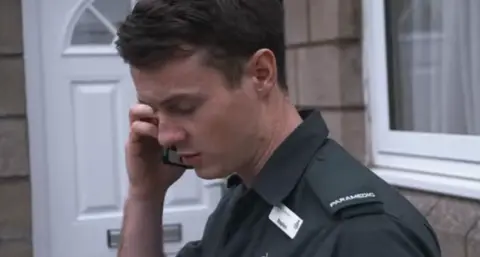 Stephen Doohan is on the phone outside of a house. He has short brown hair and looks down. He is wearing a green NHS paramedic uniform