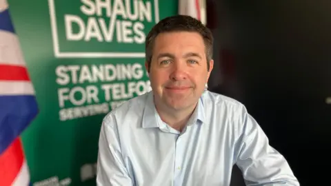 A dark-haired man wearing a pale blue shirt, sitting in front of a green and white sign that says "Shaun Davies, standing up for Telford". Two flags, including the Union flag, can be seen eighter side of the banner.