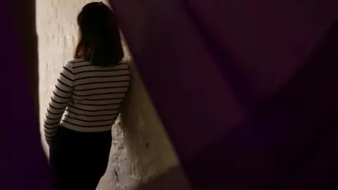 Reuters A woman in a white top with black stripes leans against the lit area of a white wall. 