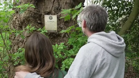 Niovi and her father look at one of the signs hung on a tree. He has her arm around her.