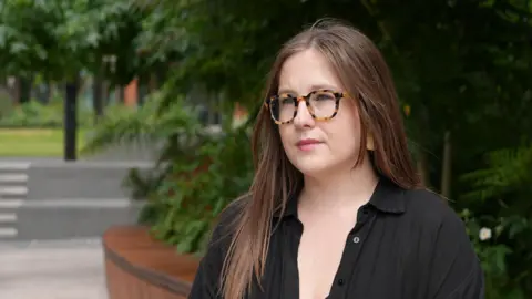 Jade Blue McCrossen-Nethercott, a woman with glasses wearing a black top, sits down looking away from the camera. There are trees behind her.