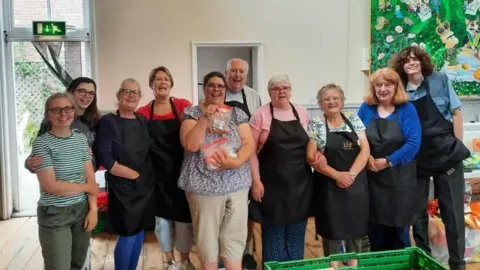 A group of people look into the camera and smile. Most of them are wearing aprons. They are stood inside a hall, with white walls behind them. In front there is a green plastic crate. 