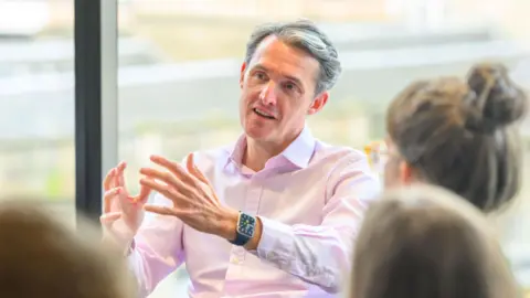 Paul Chappells/NatWest Group Paul Thwaite has short grey hair and wears a pink shirt. He gestures with his open hands facing each other and he appears to be wearing an Apple watch. He sits in front of a floor-to-ceiling window.