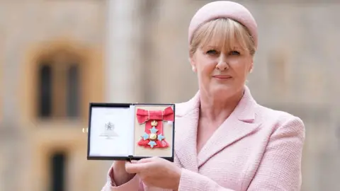 The media in the middle of a snapshot of Sarah Lancashire holds her CBE medal. She wears a pink hat.
