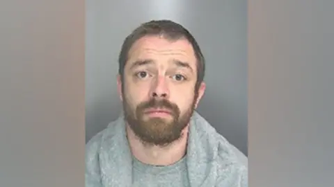 Cambs Police Police custody shot of Adam McDonnell. He is looking at the camera and has brown eyes, short brown hair and a beard and moustache. He is wearing a grey T-shirt and a grey top with a shawl collar