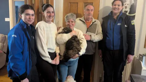 Five people stood in a line inside a house, with the woman in the middle cuddling a cat