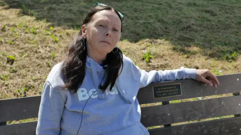 West Mercia Police A woman with long dark hair in pigtails, wearing a grey Bench hoodie, sits on a wooden bench. A plaque on the bench reads: "In loving memory of Christian Bagley - 3.6.1985-21.6.2015".