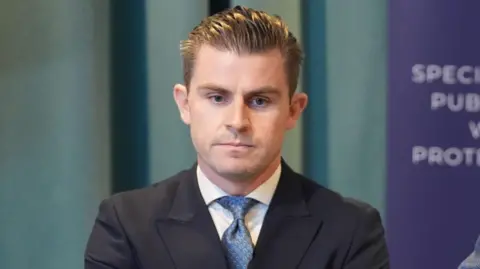 PA Media Darragh Mackin wearing a black suit jacket, white shirt and blue patterned tie. 