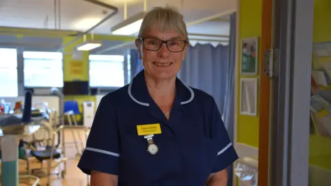 Image shows Dawn Wintle smiling into the camera with a nursing ward behind her and wearing her uniform. She wears black glasses with her hair pulled back.